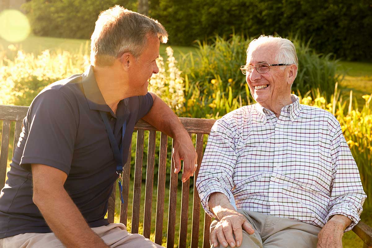 Older man sitting on outdoor bench with middle-aged son; smiling at each other