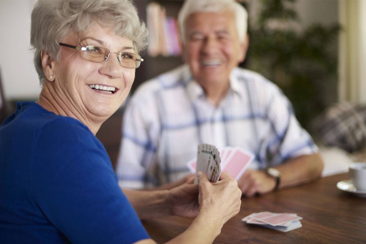 Older woman smiling of shoulder, playing cards at table with older man, smiling