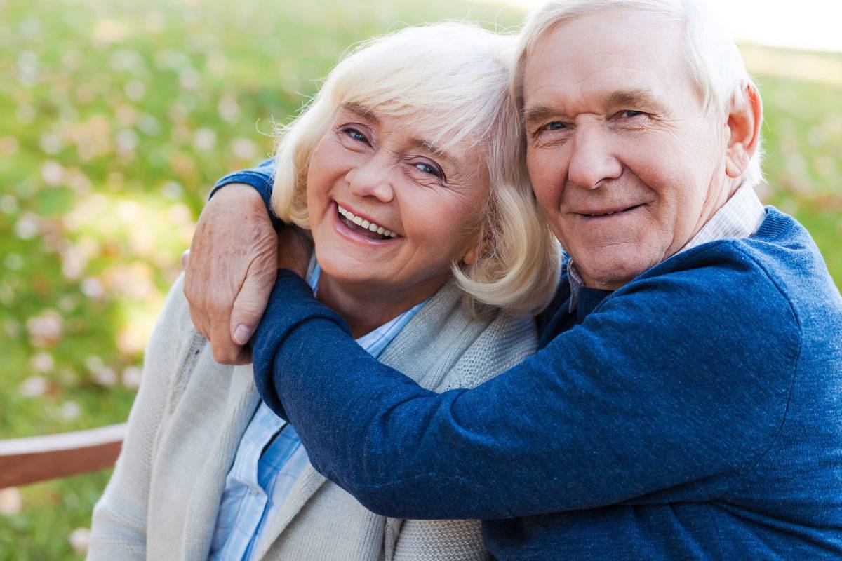 Older husband hugging with arms on wife's shoulders, sitting on bench, both smiling