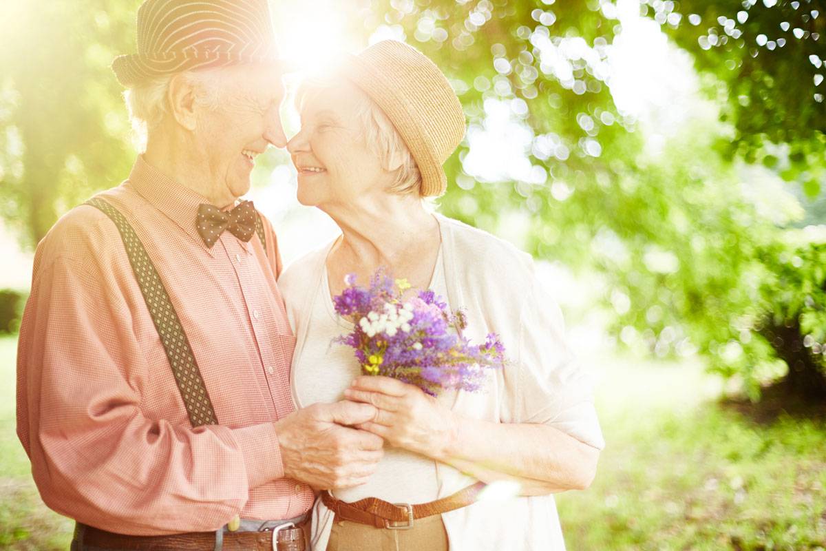 Older couple dressed up, outside, smiling to each other; woman holding flowers