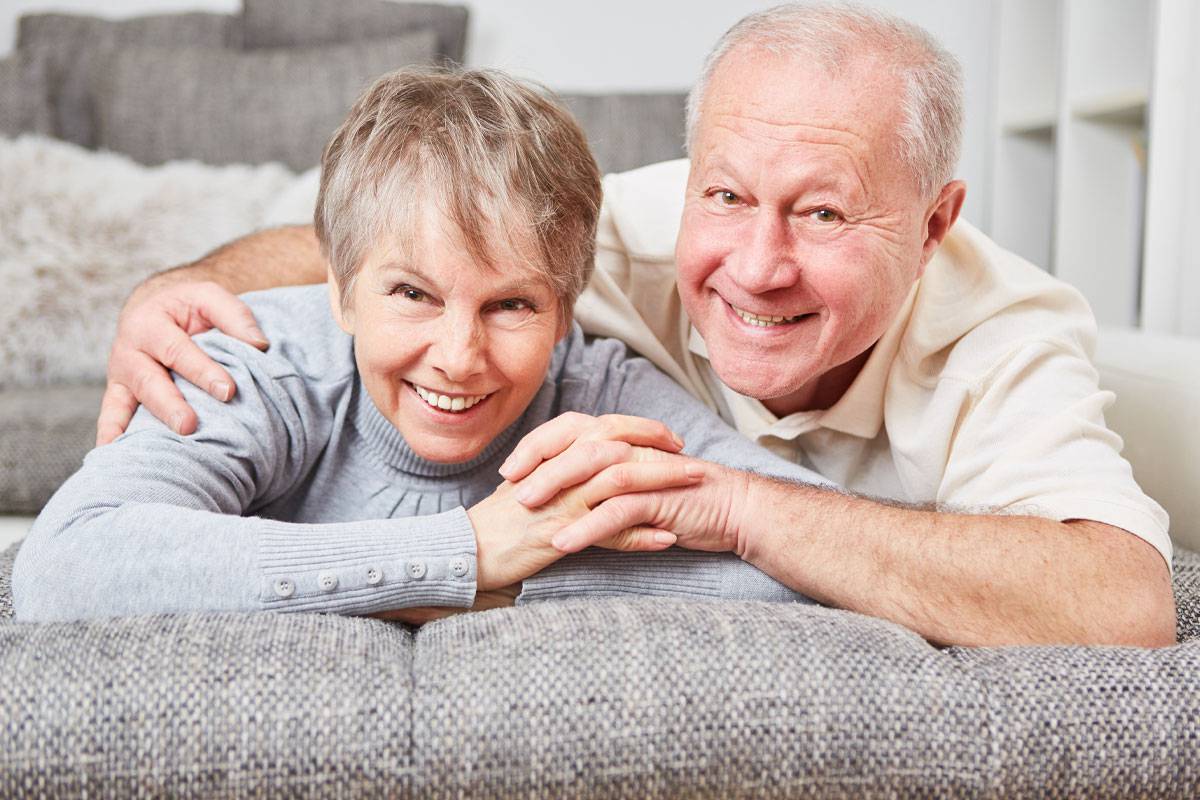 Older couple leaning over back of couch, smiling and holding hands