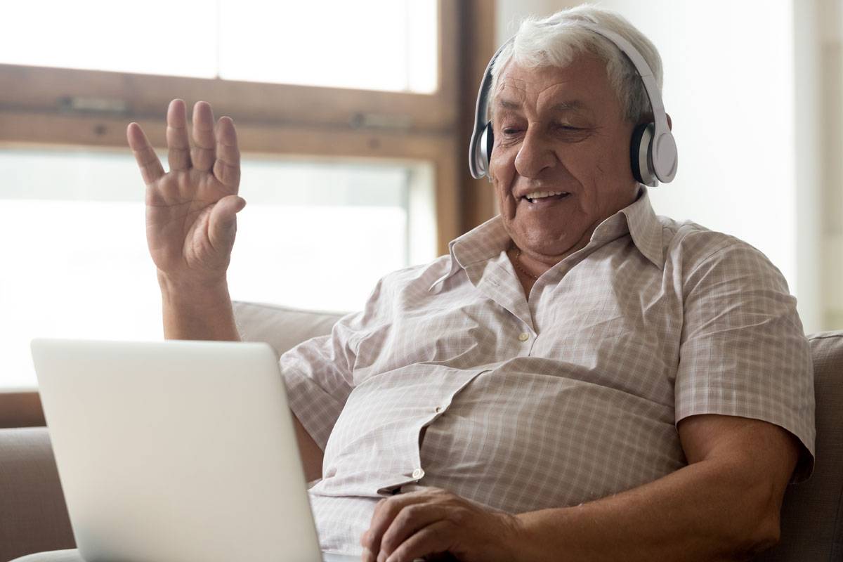 Older man wearing headphones, waving at laptop computer on his lap, sitting on couch