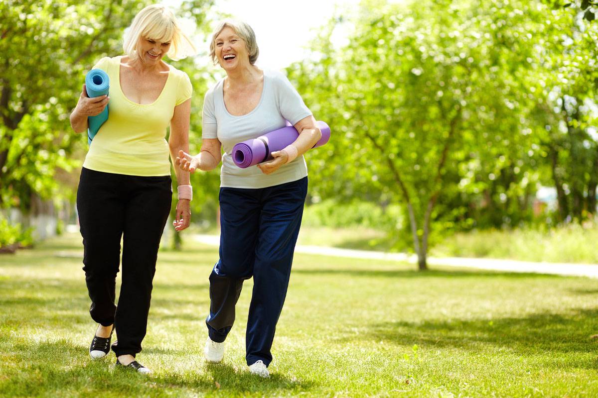 Two older woman smiling, laughing, walking outside, carrying yoga mats