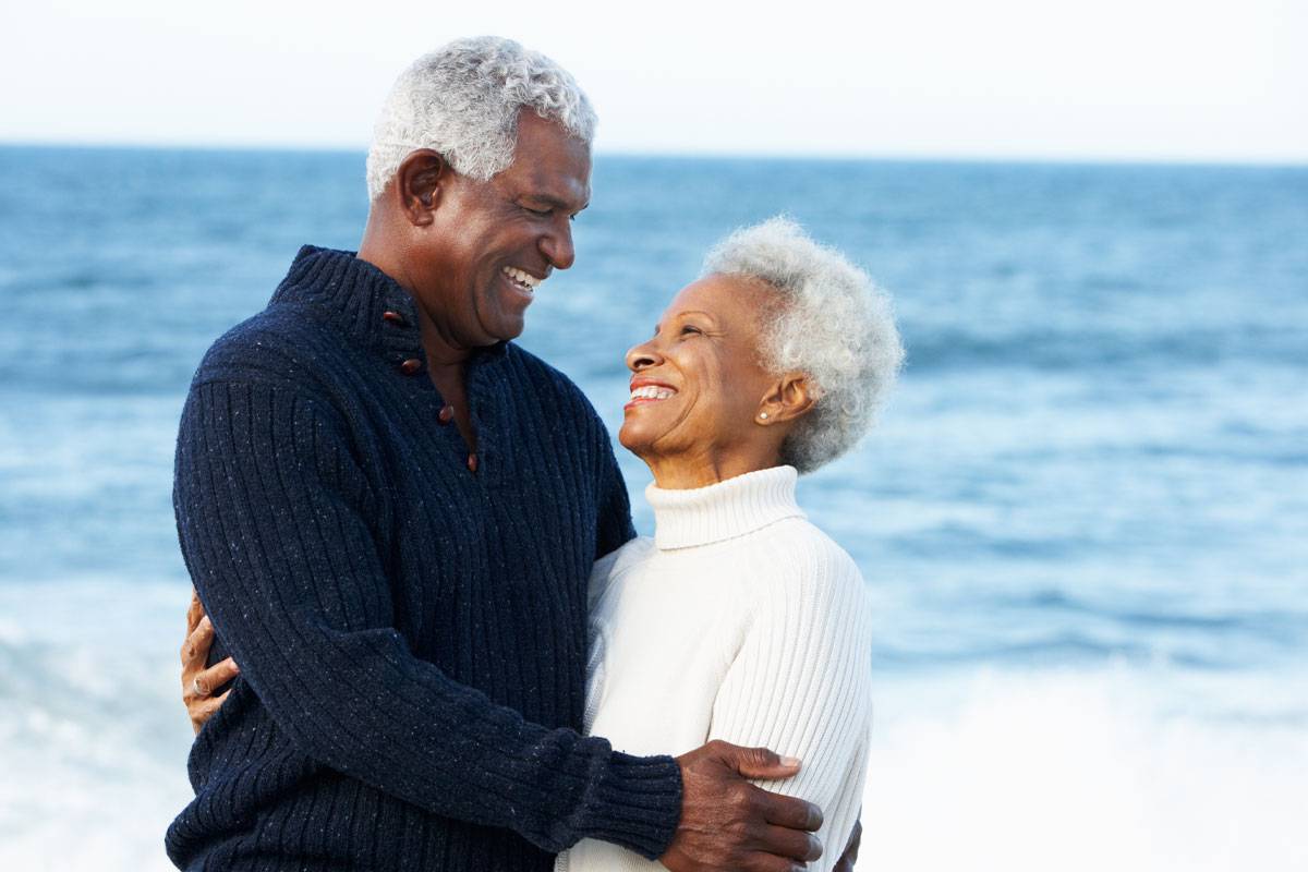Older African couple smiling at each other, holding each other, on the beach