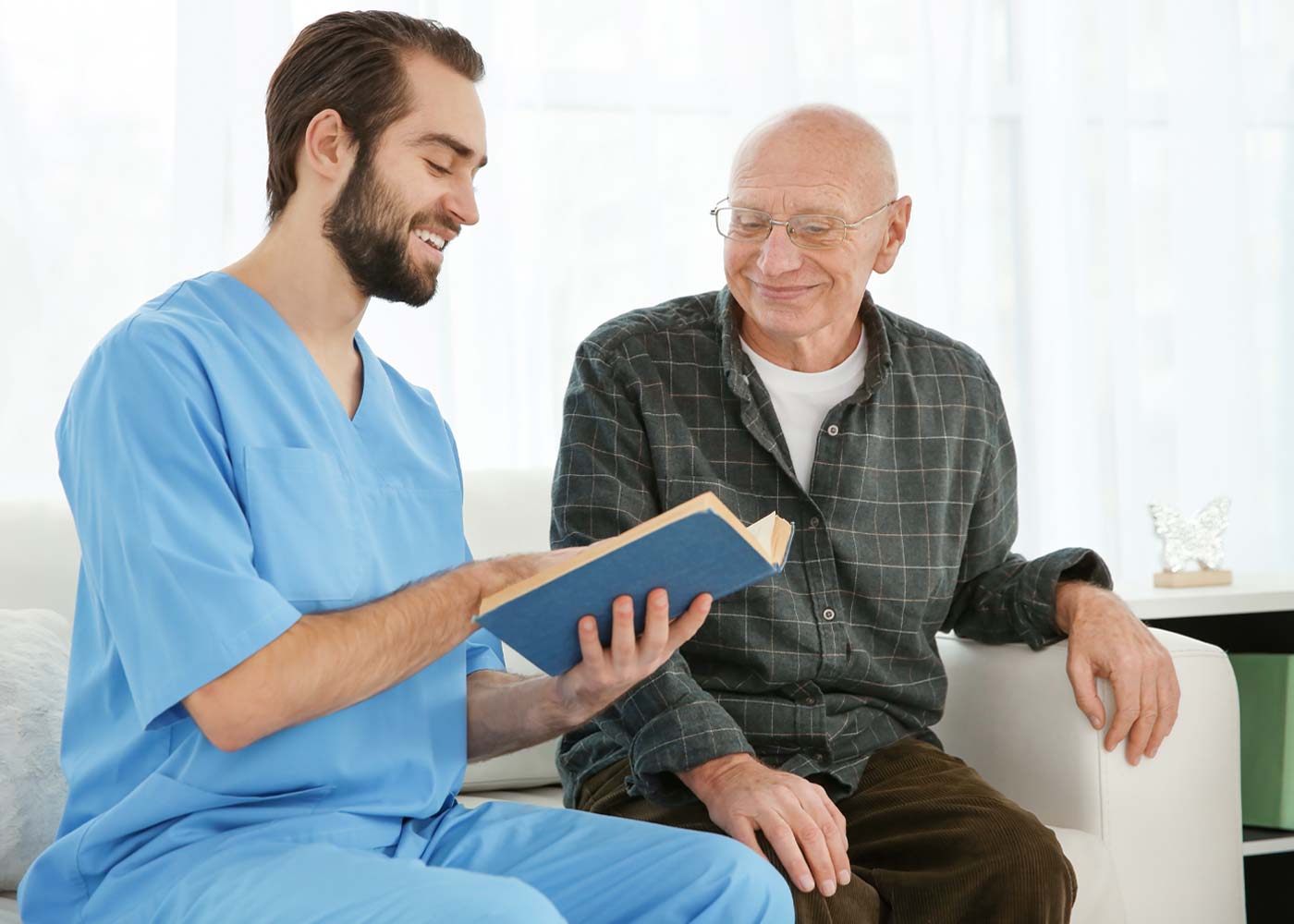 Male caregiver reading book to older man, sitting on couch