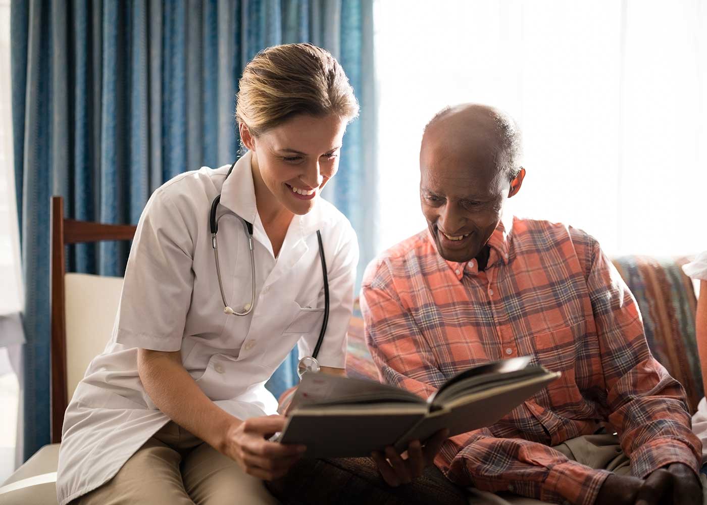 Caregiver and Older Male Smiling, Looking at Book-Memory Care Community