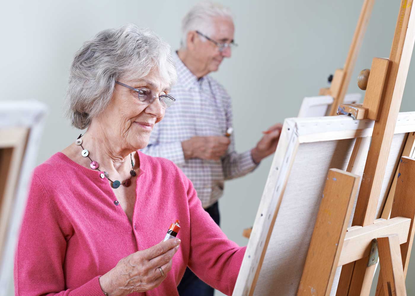 Older female in foreground, smiling at canvas on easel; older male in background painting on canvas