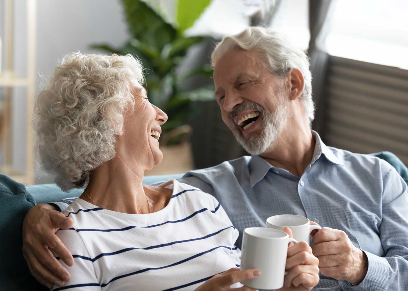 Older couple laughing together, holding mugs on couch