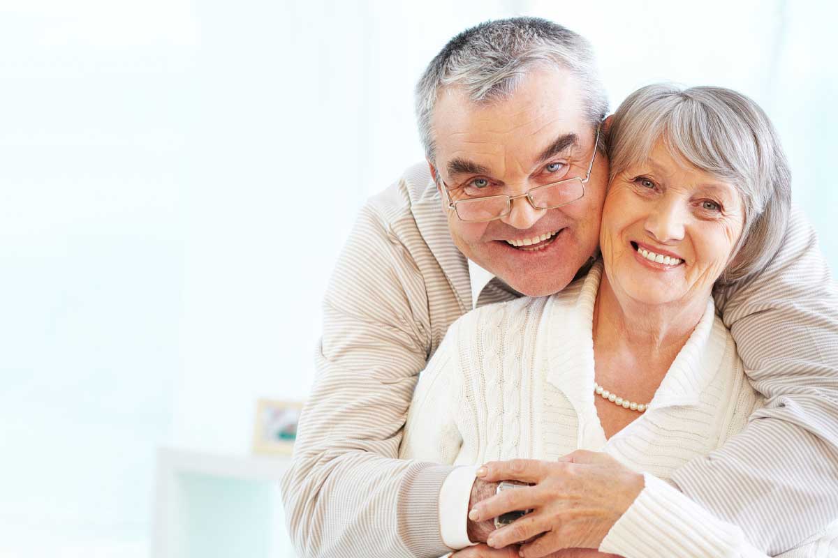 Older husband with head over wife's shoulder, both smiling