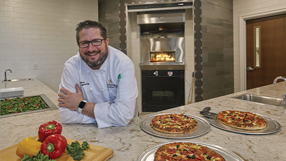 Community Chef smiling, leaning on counter with pizzas displayed-The Chef's Studio at HarborChase Senior Living