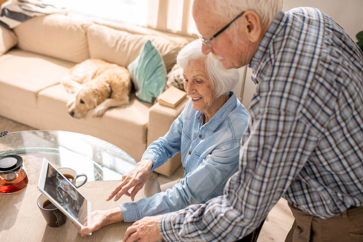 Older couple at table, looking at photos on tablet, dog on couch in background-Senior socialization and building relationships