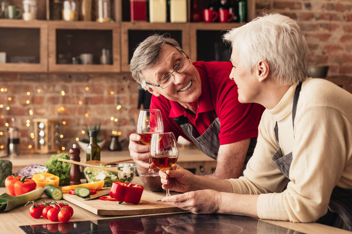Older couple leaning on kitchen counter, clinking wine glasses, cutting vegetables-Heart healthy meals