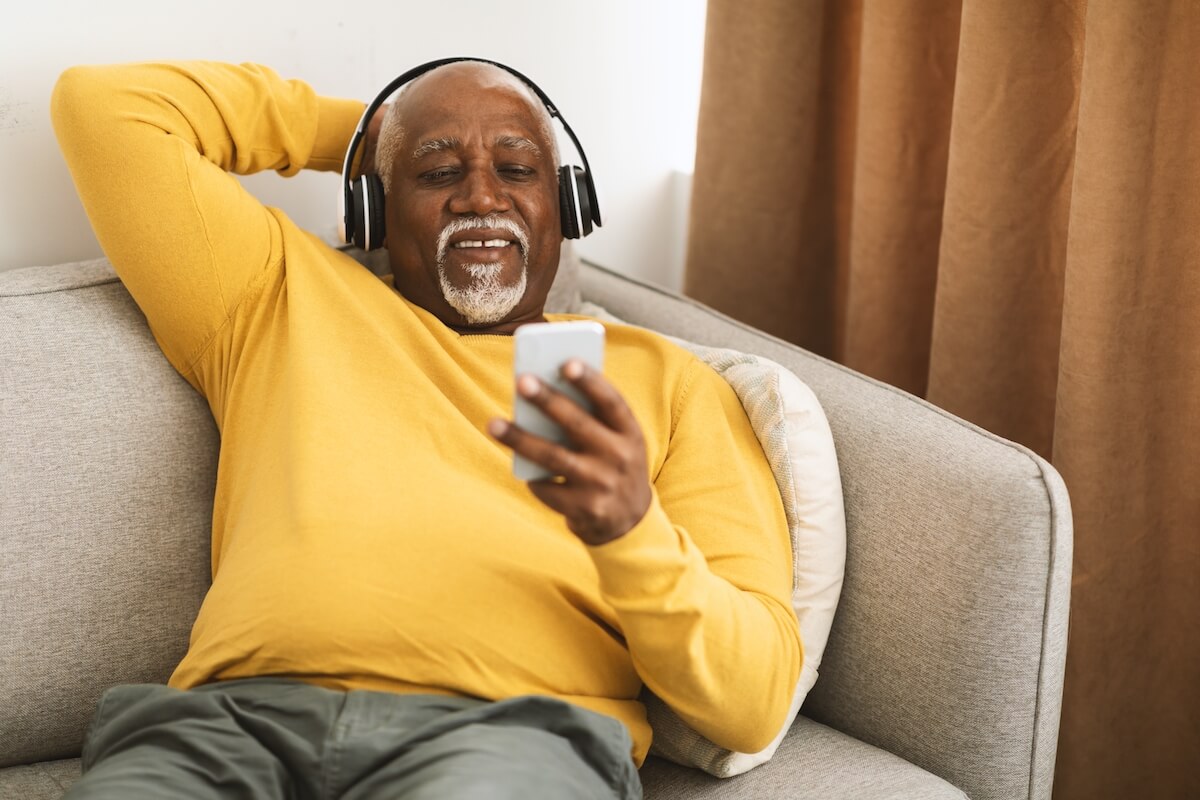 Older man sitting on sofa, wearing headphones looking at smartphone-Best Podcasts for Seniors to Listen to in 2022