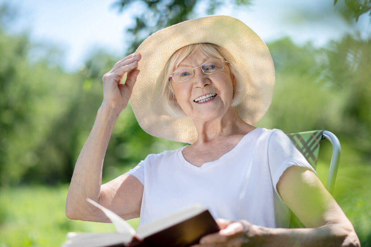 Older woman sitting outside reading, wearing large hat-Summer Safety Tips