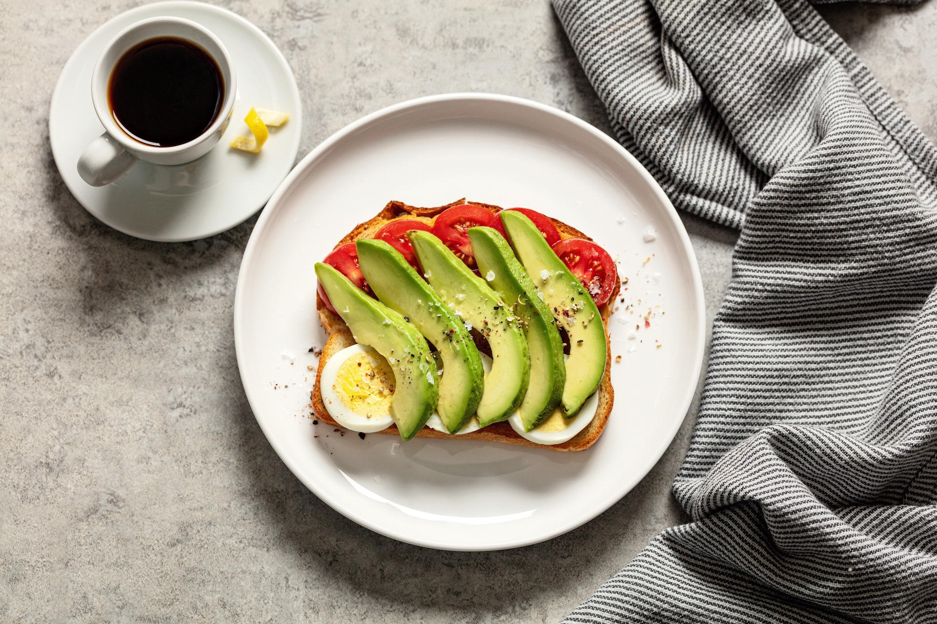 Avocado, tomato, and egg sliced on piece of toast; cup of coffee next to plate