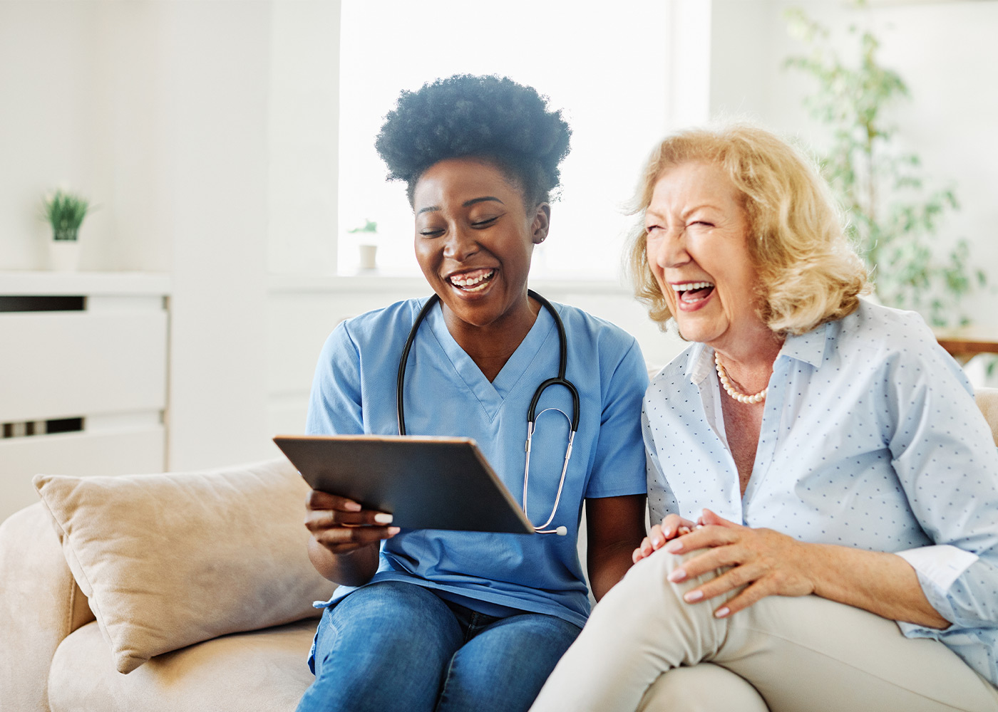 Caregiver holding tablet; sitting next to older woman; both laughing and smiling