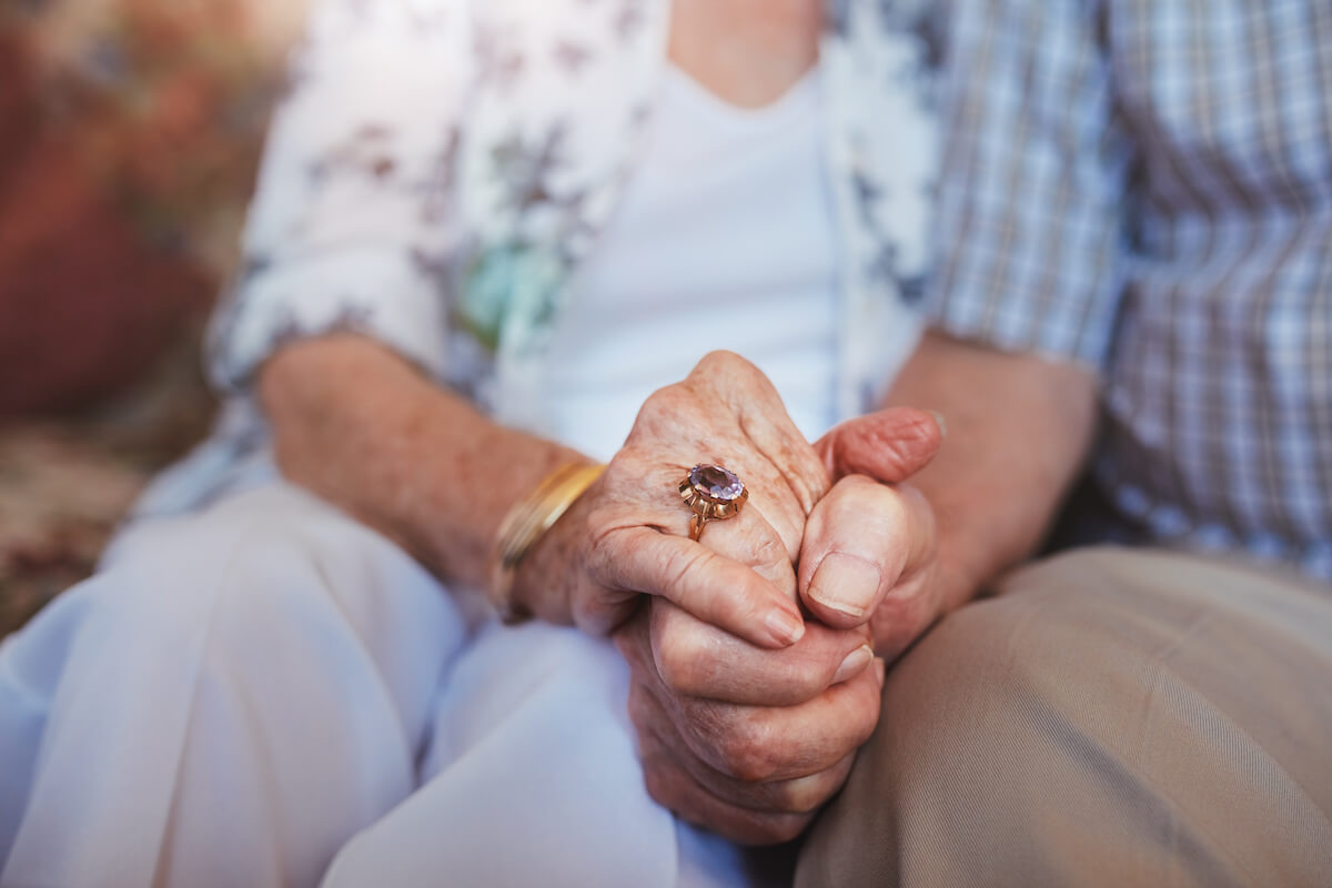 Cropped shot of elderly couple holding hands while sitting together at home. Focus on hands